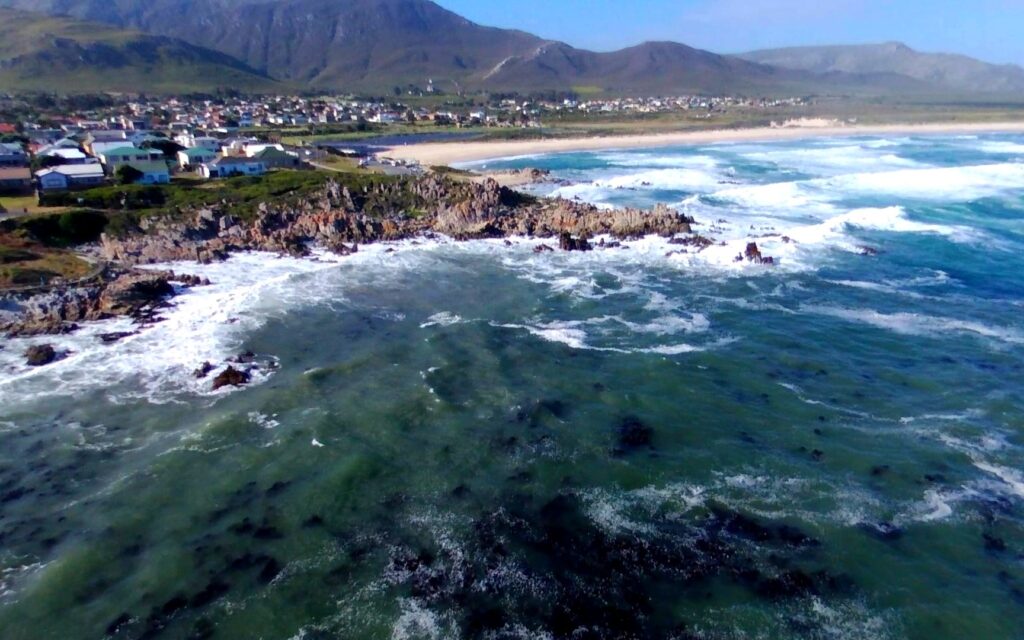 Felsküste und Brandung bei Hermanus, dahinter Strand, Ort und Berge